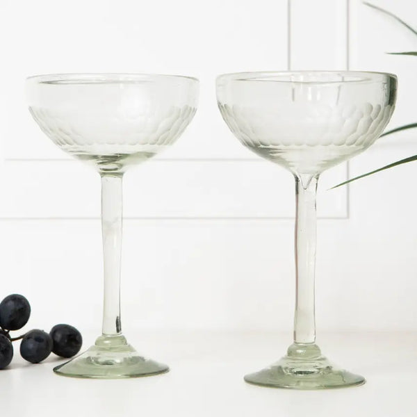 Two clear glass coupe glasses on a white background with grapes and leaves.