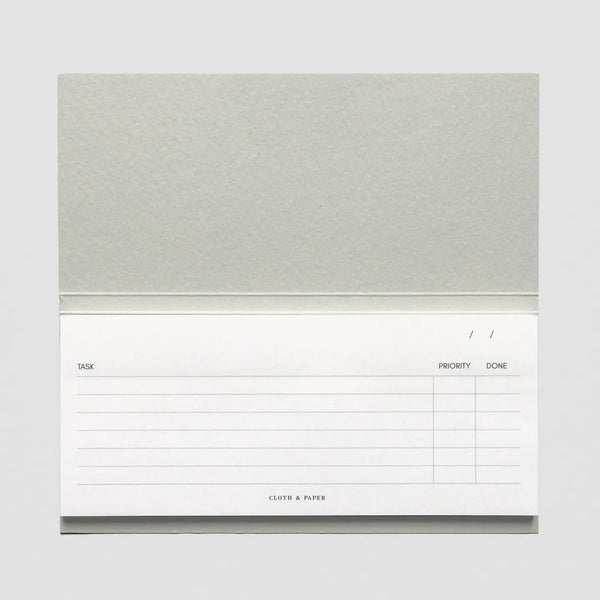 A single-page sticky note pad with a textured light gray cover and a white lined writing page showing columns for task, priority, and done, on a light gray background.