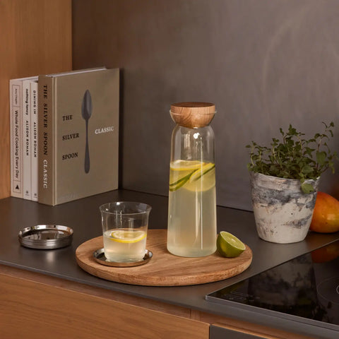 A clear glass carafe with a light wood lid sits on a round wooden tray on a dark countertop, alongside a small glass of lemon water, a metal lid, books, a potted plant, and an orange in a kitchen setting.