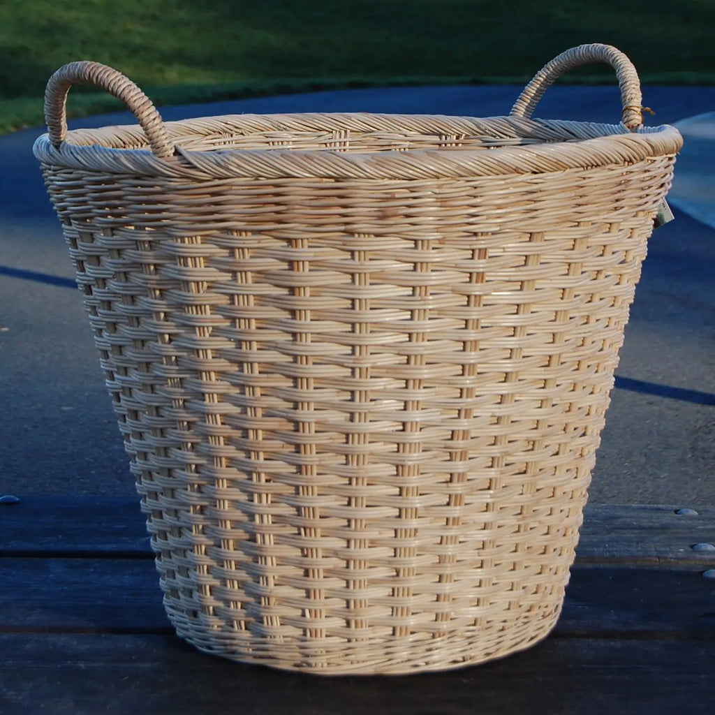 A natural woven wicker basket with two loop handles, sitting on a dark wooden surface outdoors.