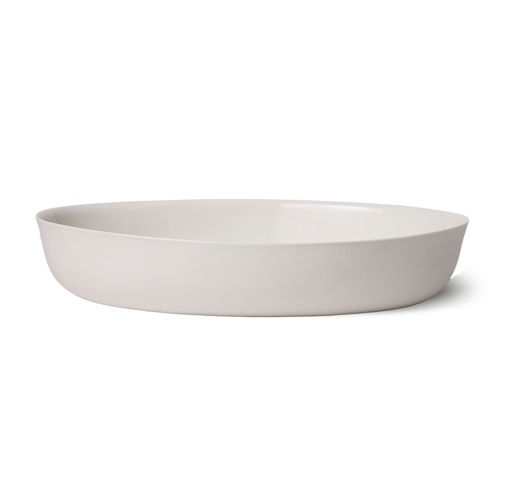 A minimalist, shallow oval ceramic bowl in a light warm gray or off-white color, shown from a side profile on a seamless white background.