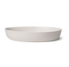 A minimalist, shallow oval ceramic bowl in a light warm gray or off-white color, shown from a side profile on a seamless white background.