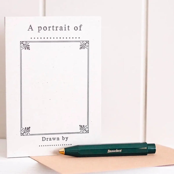 A white portrait paper/card with decorative corners and the text 'A portrait of' and 'Drawn by' stands upright next to a dark green pen on a beige paper sheet against a white paneled background.