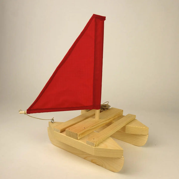 A small wooden toy sailboat with a bright red fabric sail and light natural wood hull, photographed against a plain light beige studio background.
