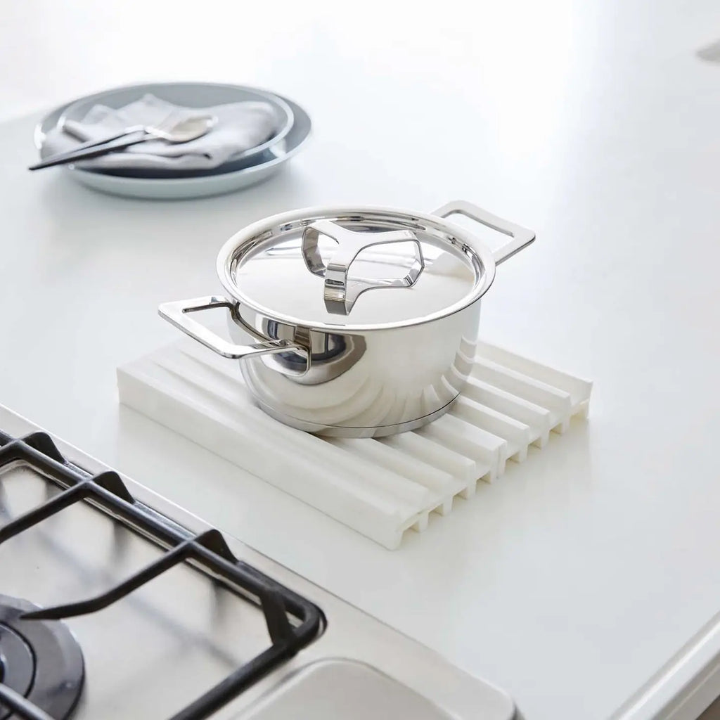 A stainless steel pot with a lid sits on a white ribbed silicone drying mat on a white kitchen countertop, with a gas stovetop burner partially visible in the foreground and a plate in the background.