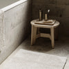 Bathroom corner with a wooden stool, bath products, and a bath mat.