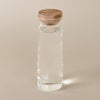 A clear glass water bottle with a round light brown wooden lid, shown on a solid beige background.