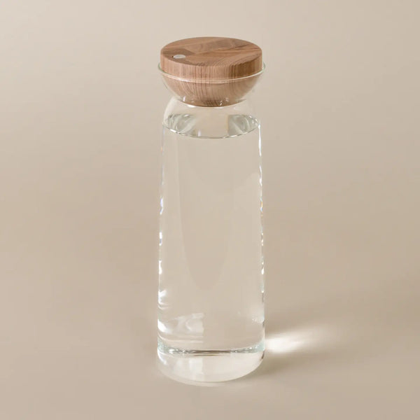 A clear glass water bottle with a round light brown wooden lid, shown on a solid beige background.