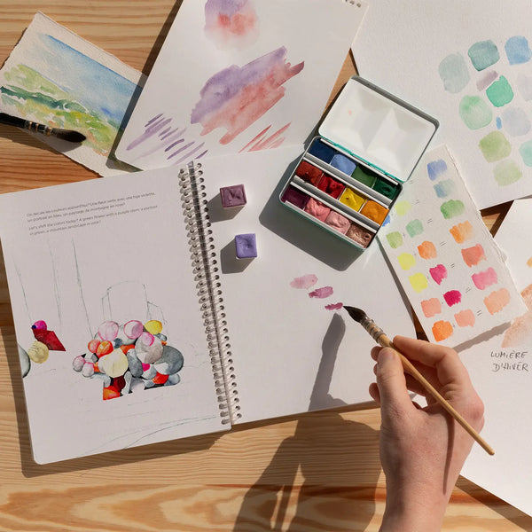 Artistic scene with watercolor paintings, a sketchbook, and a hand holding a brush on a wooden surface.