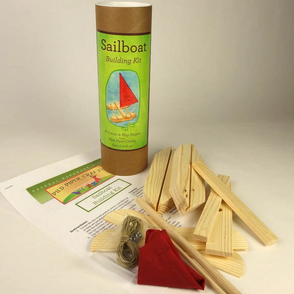 Sailboat Building Kit contents displayed on a white surface, including a brown cardboard tube labeled 'Sailboat Building Kit,' wooden splines, dowels, a red fabric sail, small metal rings, and printed instruction sheets.