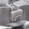 A white plastic sink-mounted sponge holder with a sponge and steel wool scrubber, hanging over a stainless steel kitchen sink.