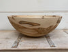 A large handcrafted wooden bowl with natural light tan wood and dark brown grain patterns, displayed on a rustic weathered wooden surface against a white paneled wall.