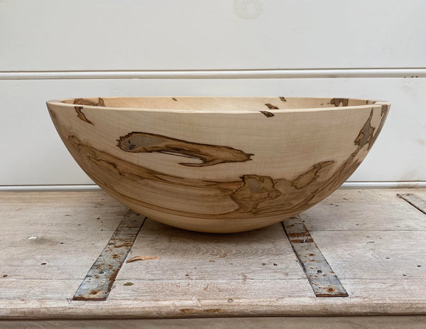 A large handcrafted wooden bowl with natural light tan wood and dark brown grain patterns, displayed on a rustic weathered wooden surface against a white paneled wall.