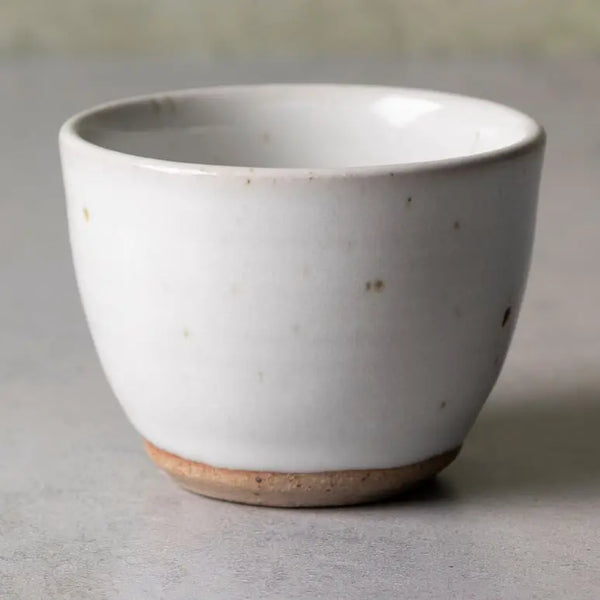 A handmade ceramic cup with a white glazed exterior and a natural, speckled tan clay base, photographed on a neutral gray surface.