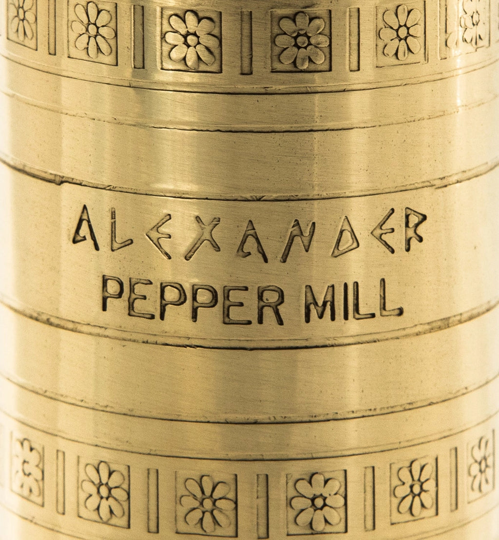 Close-up of a cylindrical brass or gold-toned metal object engraved with the words 'ALEXANDER PEPPER MILL' and decorated with floral motifs.