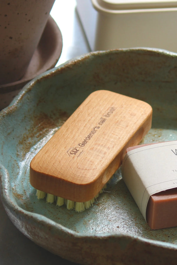 A wooden garden brush with light green bristles and a white paper sleeve, resting in a rustic teal ceramic dish next to a bar of soap.
