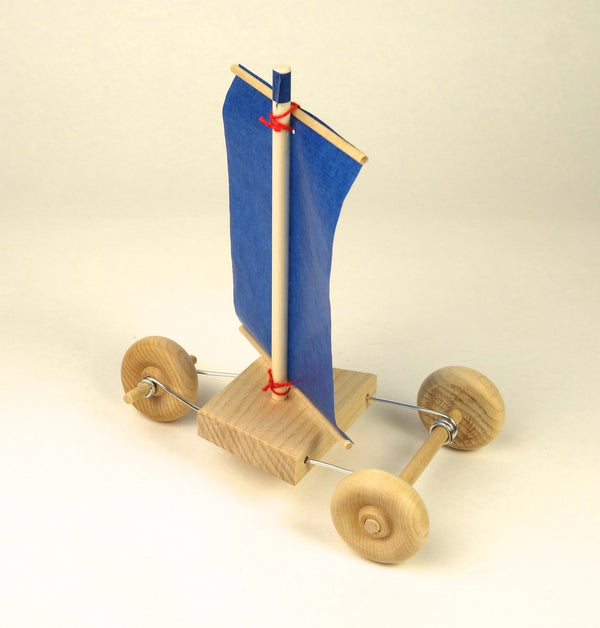 A small wooden toy sailboat with a blue fabric sail, natural wood hull and wheels, and thin metal axles, photographed on a plain light beige studio background.