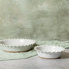 Two round white ceramic pie bowls with scalloped edges and a speckled finish, displayed on a light gray surface with a pale green cloth and textured wall background.
