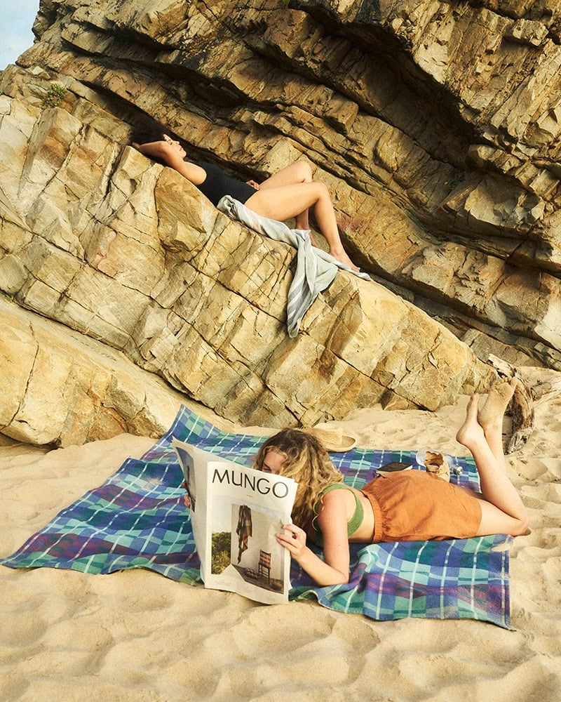 Two people on a rocky beach, one reading a magazine and the other sunbathing.