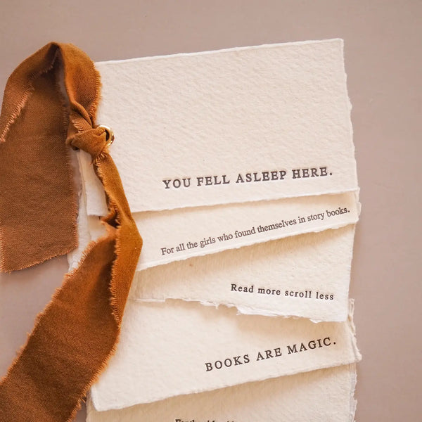 A close-up of a handmade layered off-white paper pages featuring printed quotes, tied with a rustic brown suede ribbon, on a neutral beige background.