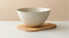 A matte off-white ceramic bowl resting on a light natural wood serving board against a warm beige background.