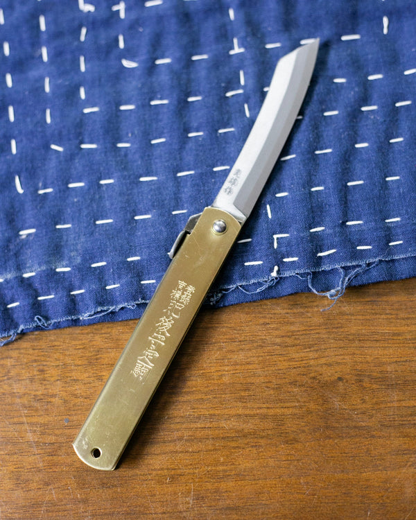 Small folding knife, brass, with Japanese lettering. Laying on a wooden table on top of a blue towel with white stitches