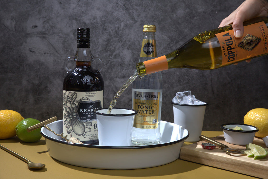 Person pouring a bottle of liquor into a glass with tonic water on a table and a serving tray.