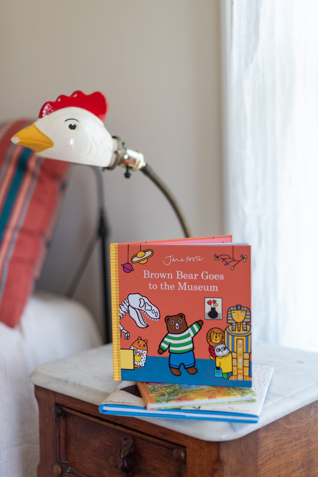 A children's picture book titled 'The Brown Bear Goes to the Museum' stands open on a small wooden table next to a decorative lamp shaped like a white rooster with a red comb and yellow beak.