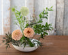 A floral arrangement in a white bowl on a wooden table, featuring two peach dahlia flowers, a pale green globe flower, and green foliage against a rustic weathered wood background.