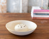 A white ceramic dish or bowl with a small  flower frog inside, set on a wooden tabletop with a blurred stack of books in the background.
