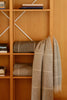 A beige and gray plaid wool blanket is draped over a wooden shelf next to folded matching blankets, with books arranged on the shelving unit.