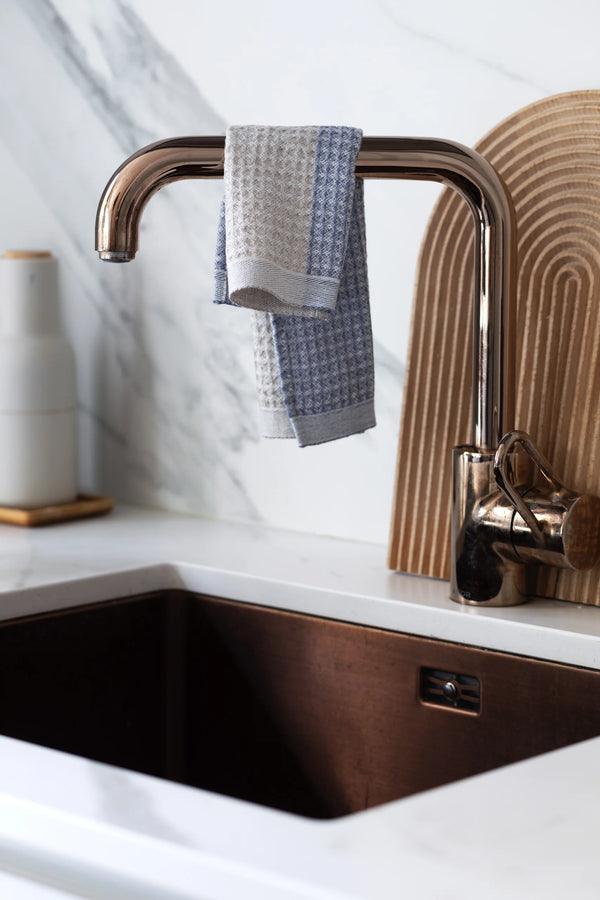 Dishtowel hanging from a kitchen faucet over a kitchen sink. A wooden cutting board behind the faucet.
