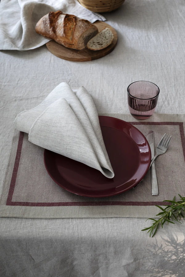A table setting with linen placemat, tableware and linen napkin. in the background a piece of bread on a wooden cutting board. 