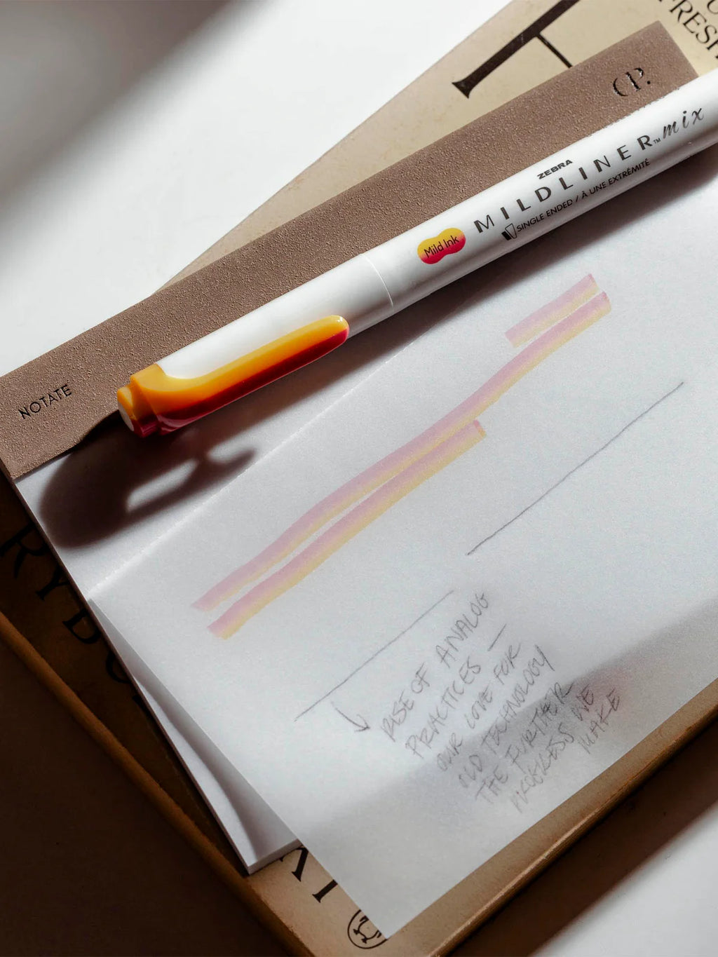 A white Mildliner gel pen with a translucent orange and yellow cap lies diagonally across a sheet of white paper. The paper shows two pink and peach-colored horizontal lines drawn with the pen, alongside handwritten notes.