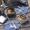 Outdoor setting with food containers, utensils, and a water bottle on a wooden surface.