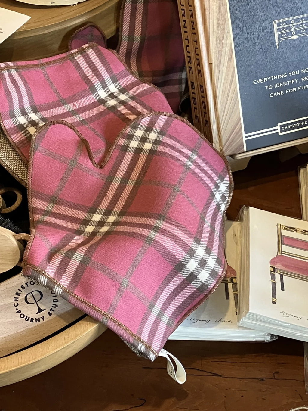 A pair of pink plaid polishing mitts with white, beige, and gray tartan lines, resting on a wooden table next to a book and other home decor items.