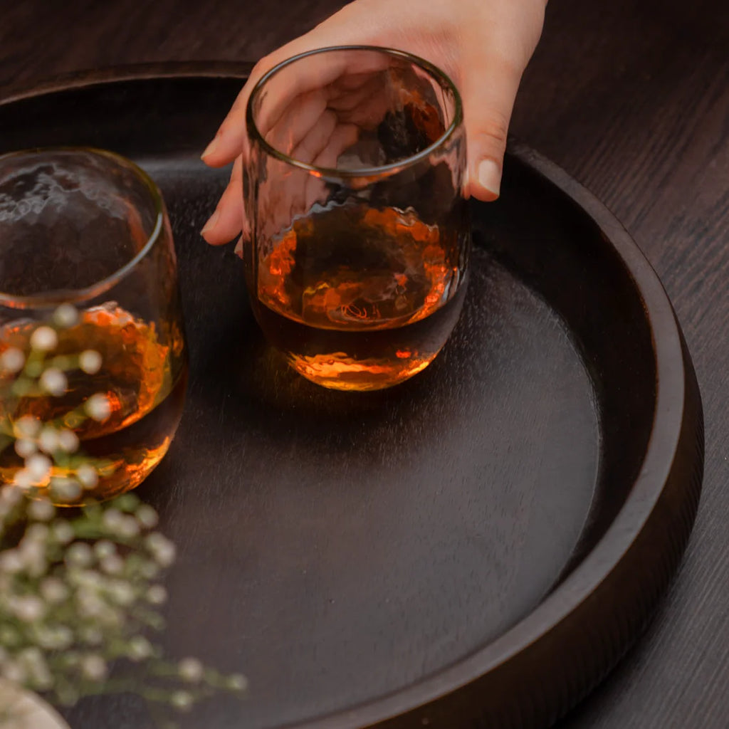 Hand holding a glass of amber liquid on a dark wooden tray with another glass and small plant.