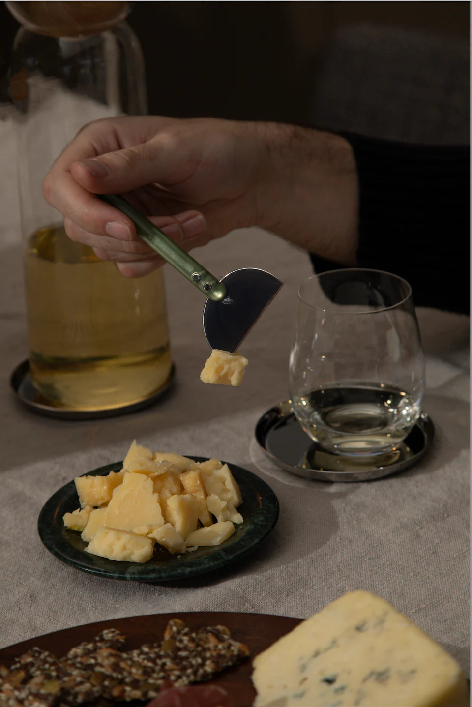 Person using a cheese knife to cut cheese on a plate with a glass of beer and another glass in the background.