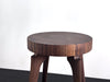 A round wooden side table with a thick, chunky top and four sculptural legs, shown against a plain white wall and dark floor.