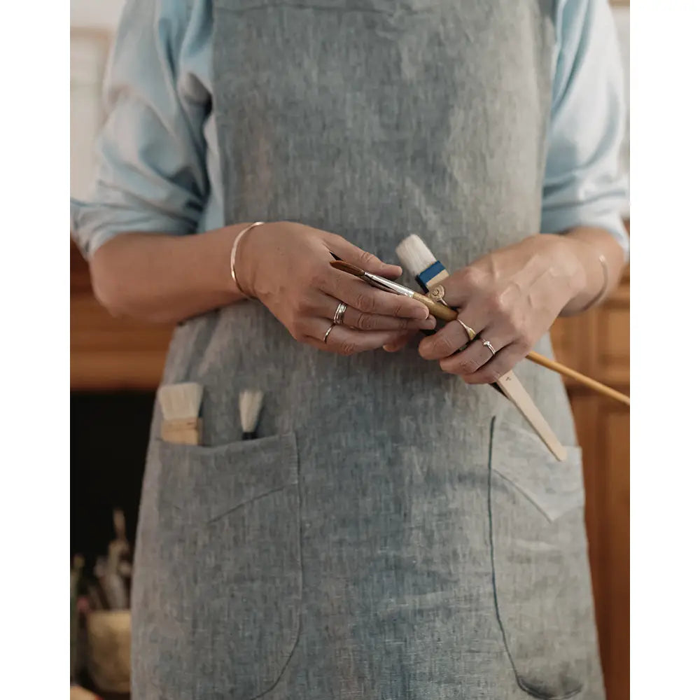 A person wearing a light blue shirt and a gray linen apron, holding a small tool in both hands. The apron has front pockets and is shown in a warm indoor setting.