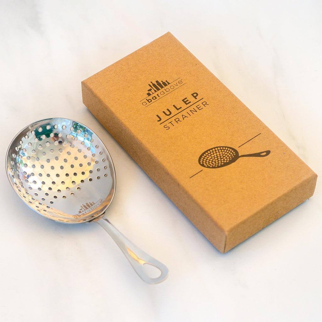 A stainless steel julep strainer with a perforated bowl and handle, shown next to its brown kraft paper packaging box on a light marble surface.