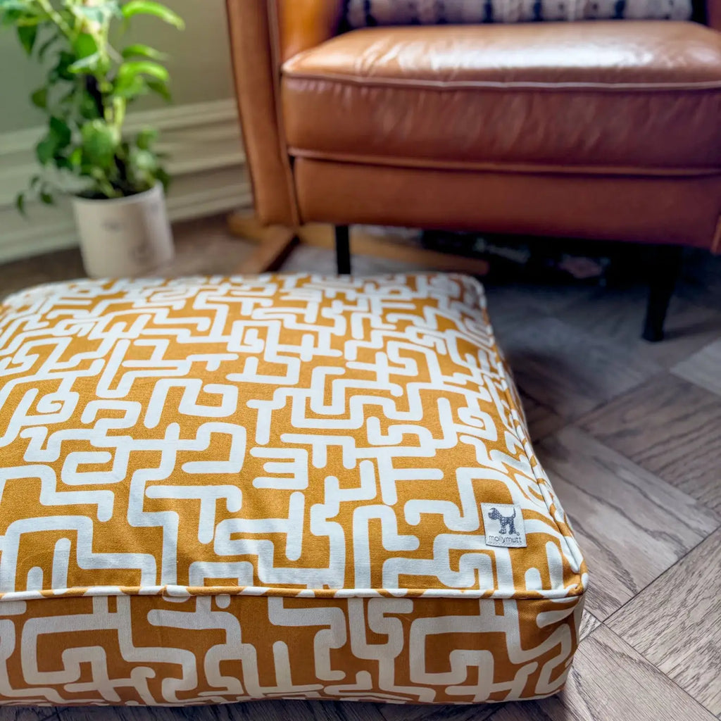 Patterned dogbed on a wooden floor with a brown leather chair and plant in the background