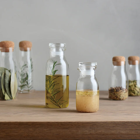 A set of heat-resistant glass dressing bottles with cork stoppers, filled with various liquids, displayed on a wooden surface.