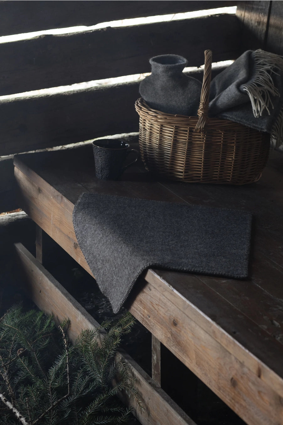 A dark charcoal gray wool seat pad is draped over a rustic wooden bench, with a matching gray throw and a wicker basket holding a dark vase in the background.