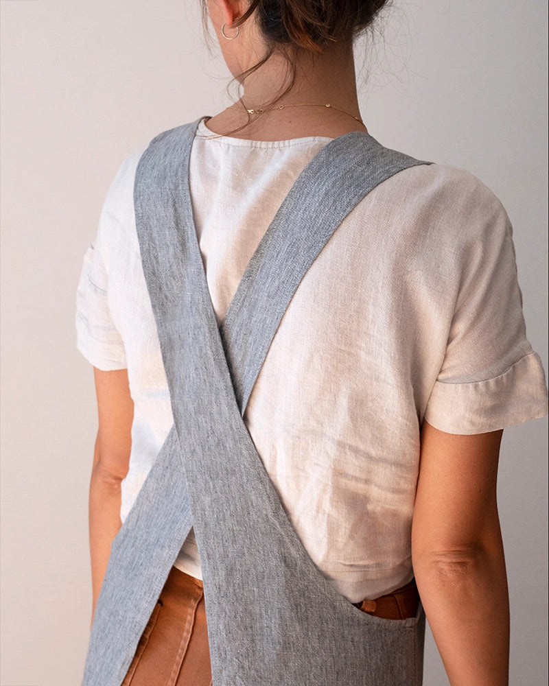 A person shown from the back wearing a light gray linen crossbody apron over a white linen t-shirt and brown leather pants, against a plain light beige background.