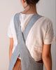 A person shown from the back wearing a light gray linen crossbody apron over a white linen t-shirt and brown leather pants, against a plain light beige background.