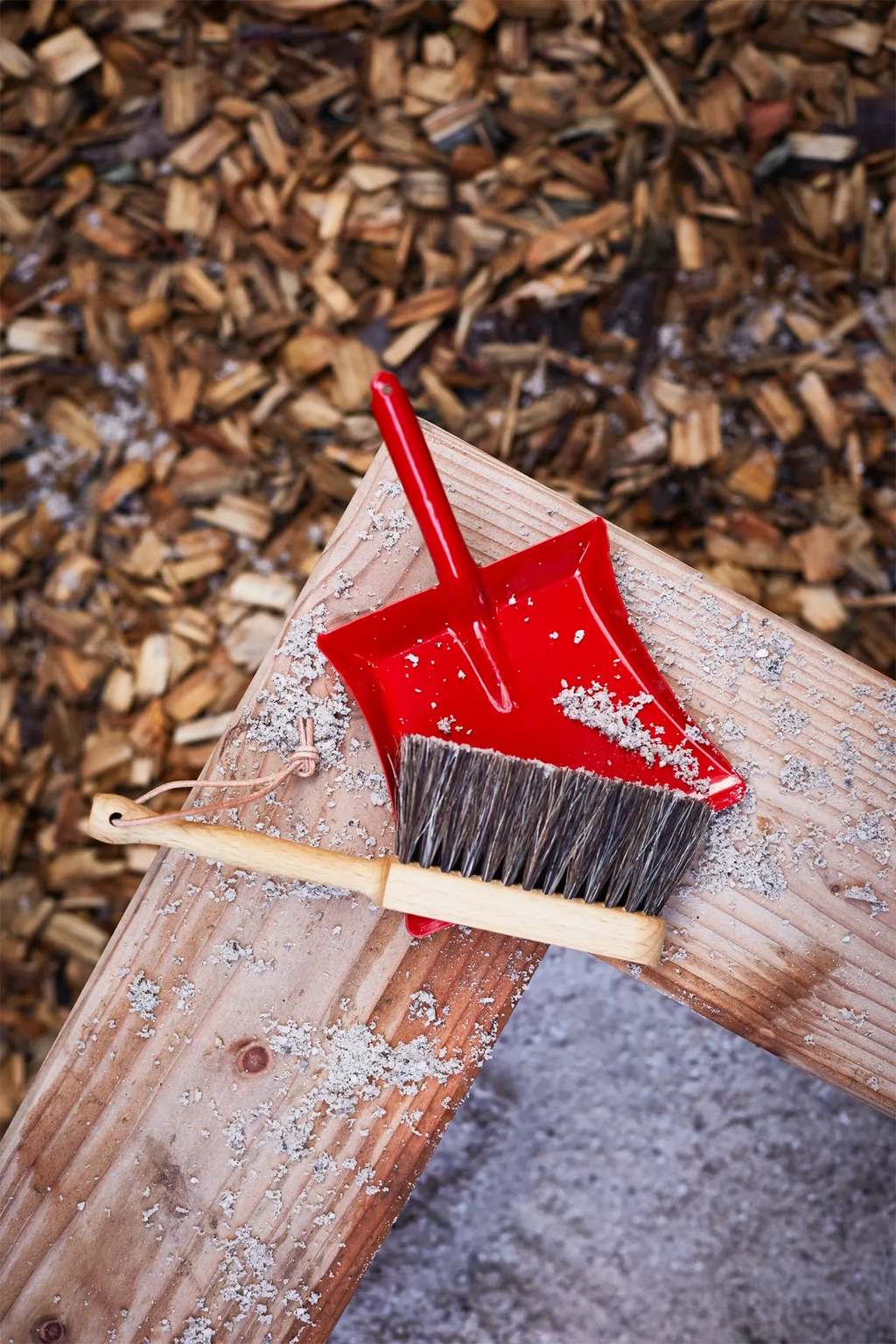 Red dustpan and broom on a wooden surface with wood chips in the background