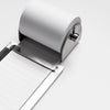 A close-up of a metallic roll of lined paper unspooling from a silver metal dispenser on a clean white background.