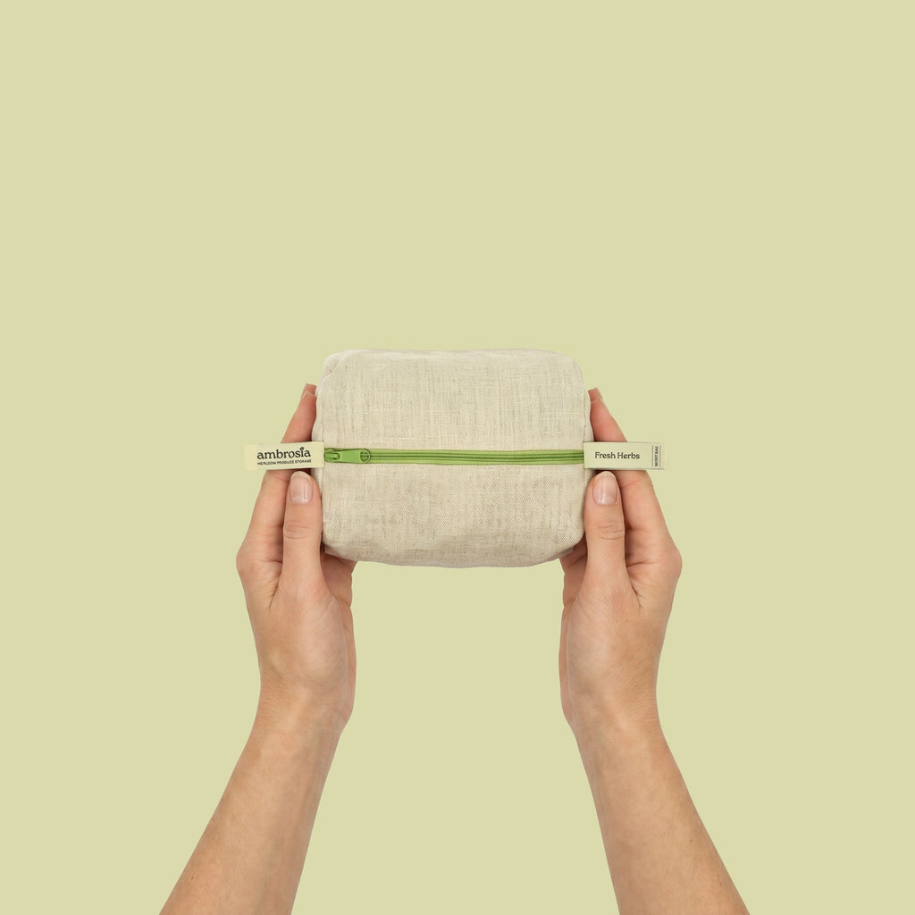 A person's hands holding a small rectangular beige linen pouch with a green band and white end-tags against a solid pale yellow-green background.