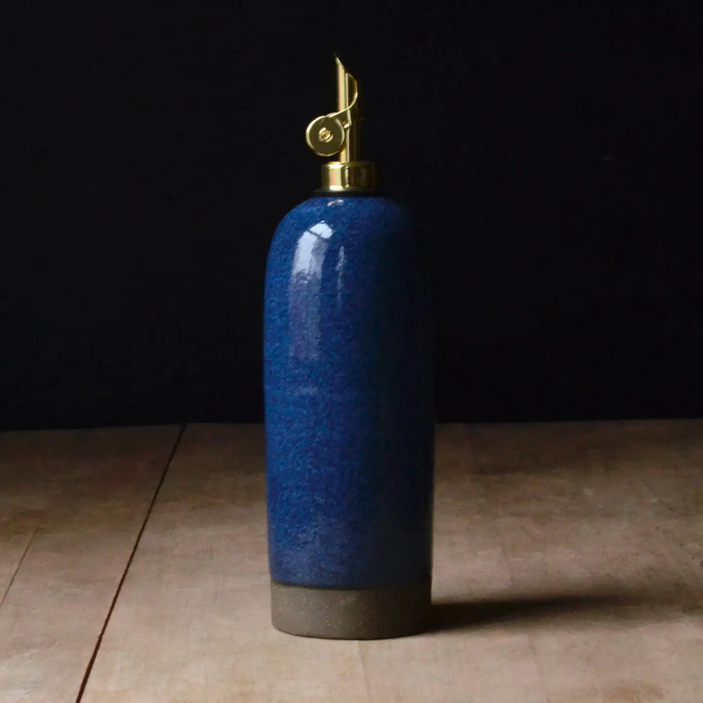 A tall cylindrical blue ceramic oil bottle with a glossy speckled glaze, a dark gray base, and a brass-toned decorative spout or cap, standing on a light wood surface against a dark background.